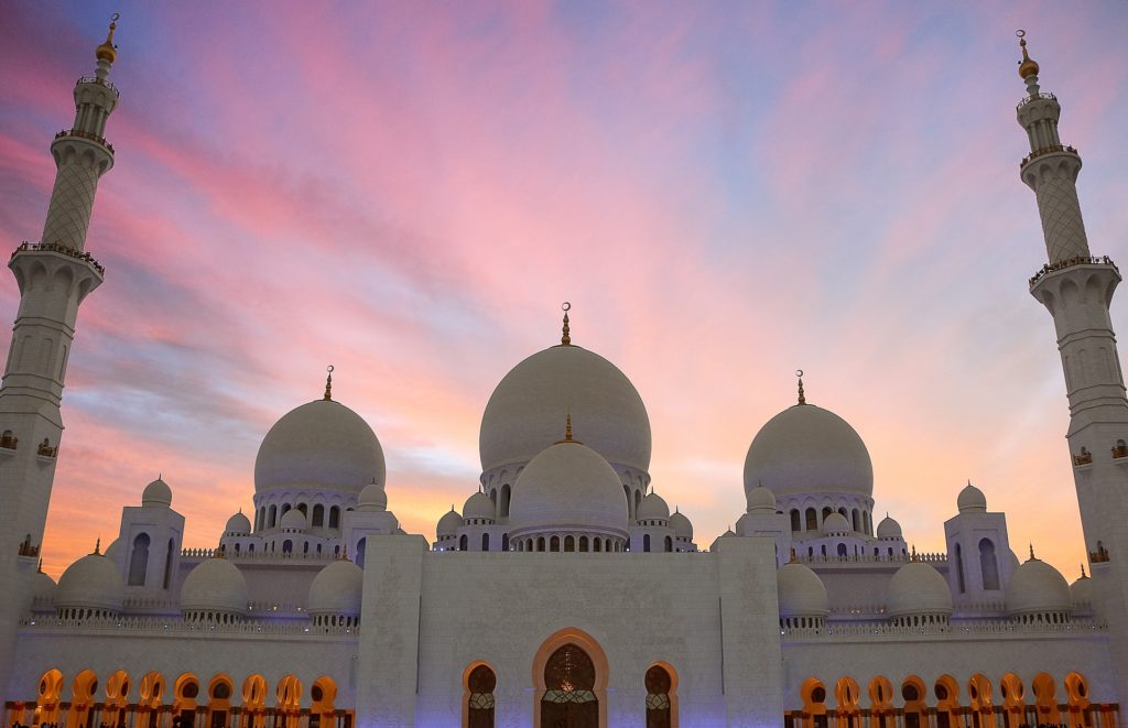 a large white building with domes and a pink and purple sky with Sheikh Zayed Mosque in the background